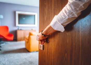 Toned man closing the door of the luxurious hotel room, man wearing a bracelet and a white shirt, closing the wooden door, nice and luxurious hotel room in the background.