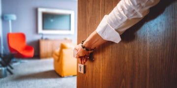 Toned man closing the door of the luxurious hotel room, man wearing a bracelet and a white shirt, closing the wooden door, nice and luxurious hotel room in the background.